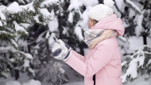 Ung kvinna klappar händerna i vantar med snö i en magisk skog i vinter, i bakgrunden på fluffigt snöiga fir trees slowmotion — Stockvideo