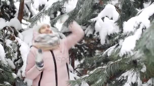 Hermosa joven mujer balanceándose y arrojando nieve de las ramas de los árboles, riendo y divirtiéndose en el bosque de invierno — Vídeos de Stock