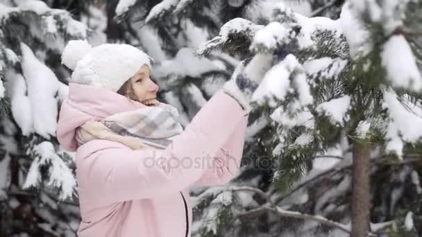 Веселая молодая женщина в зимнем лесу стряхивает снег с еловых ветвей и улыбается замедленным движением — стоковое видео
