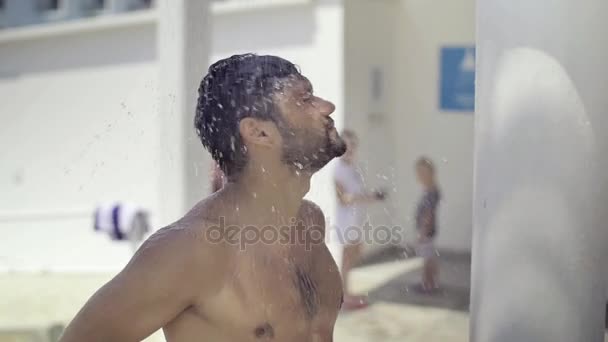 Sportovní mladý muž bere sprchu na pláži, dotkne jeho vlasy, kroutí hlavou, šplouchání vody v různých směrech. Zpomalený pohyb — Stock video