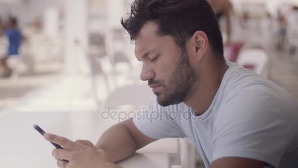 Porträtt av en söt ung man med en smartphone medan du sitter på ett café i sommar i bra väder — Stockvideo