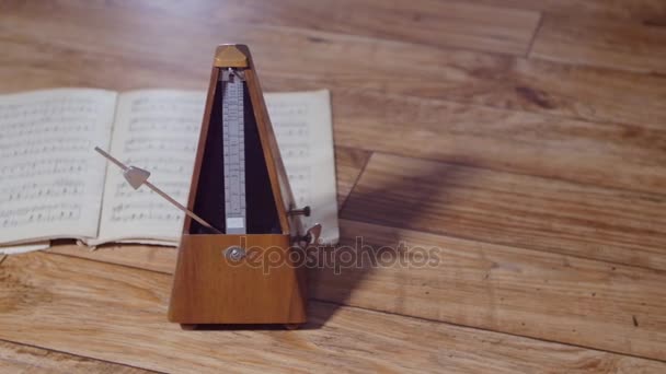 Panning shot vintage metronome with a silver pendulum beats a slow rhythm, in the background an open musical book — Stock Video