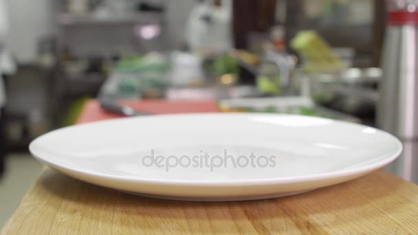 Lege schone witte plaat staat op een houten tafel. Plaat bereid te dienen van een maaltijd in een restaurant of café. Op de achtergrond typisch catering keuken — Stockvideo