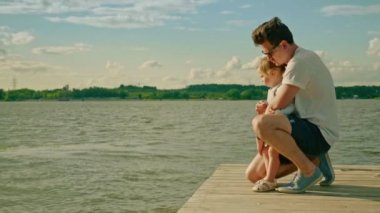 Güneş gözlüğü takan bir adam kızıyla birlikte tahta iskelede oturuyor.