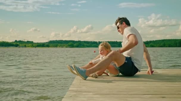 Hombre guapo divertida hija sentarse en el muelle del río a la luz del sol — Vídeos de Stock