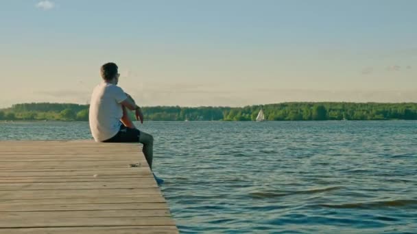 Ontspannen jongeman zit op houten pier rand kijken rivier — Stockvideo