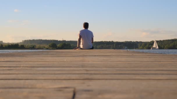 Muž v bílém tričku sedí na kraji mola a dívá se na řeku — Stock video