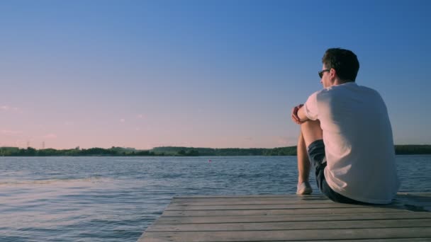 Joven se sienta en el borde del muelle de madera contra el río increíble — Vídeos de Stock