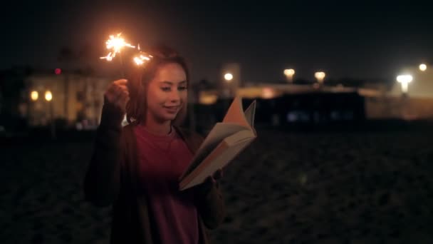 Тысячелетняя женщина читает книгу, держа в руках искры на вечернем пляже — стоковое видео