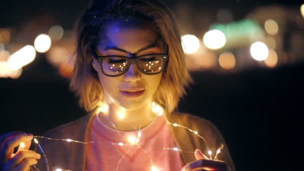 Trendy millenial girl in glasses holding garland standing at night outdoors medium close up — Stok video