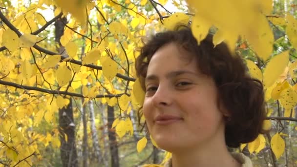 年轻漂亮黑发的女孩望着镜头微笑。在黄色的树叶的树. — 图库视频影像