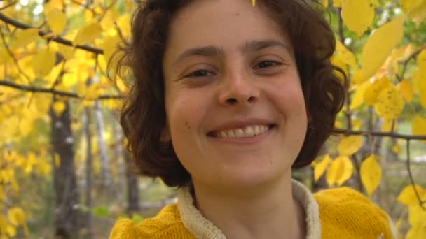 Belle fille regardant la caméra sourire. Parmi les feuilles jaunes de l'arbre . — Video