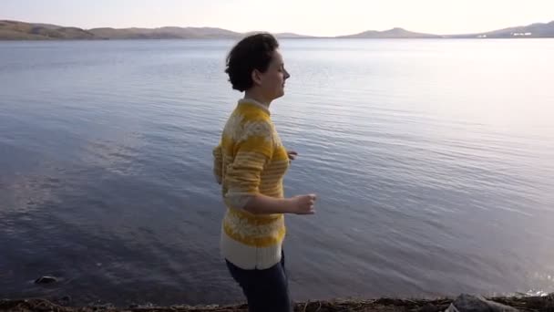 Młoda dziewczyna biegnie wzdłuż brzegu jeziora na tle wody — Wideo stockowe