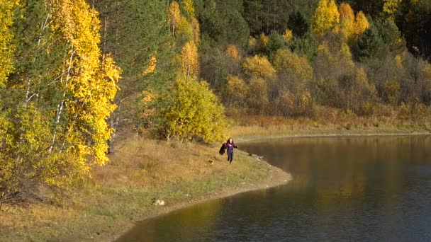 Chica corre a lo largo de la orilla de un bosque lago otoño soleado Day.small perro siguiente. Moción lenta — Vídeos de Stock