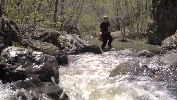 Der junge Mann springt in Zeitlupe über einen Waldbach. Vorfrühling ist ein sonniger Tag, ein großer Wasserstrom. — Stockvideo