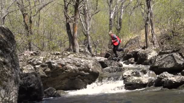 A fiatalember szőke lassított ugrik egy erdei patak felett a sziklák. Kora tavasszal egy napsütéses napon, egy nagy víz patak. — Stock videók
