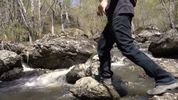 A fiatalember szőke lassított ugrik egy erdei patak felett a sziklák. Kora tavasszal egy napsütéses napon, egy nagy víz patak. — Stock videók