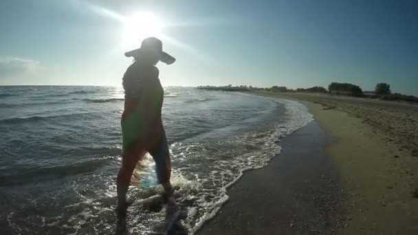 Mulher de chapéu andando ao longo do surf ao pôr do sol — Vídeo de Stock
