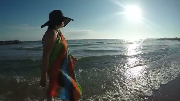 Mujer con sombrero caminando a lo largo del oleaje al atardecer — Vídeos de Stock