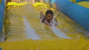 Neşeli çocuk parkta köpüklü sarı bir kayağa biniyor.