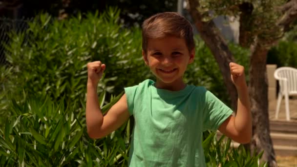 Счастливый мальчик в зеленой футболке показывает мышцы — стоковое видео