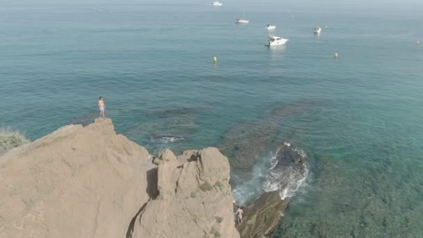 Disparos aéreos de un joven parado en un acantilado sobre un mar despejado — Vídeo de stock