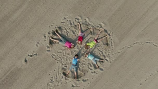 Five children lie on the sand in the shape of a star and wave their hands — ストック動画