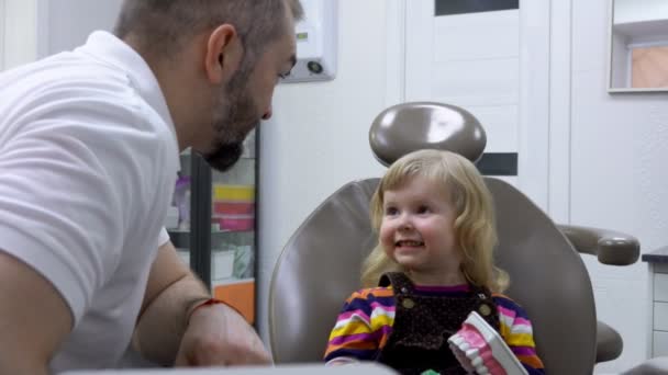 Dentist talks to a little cute girl about teeth cleaning with a teeth brush — ストック動画