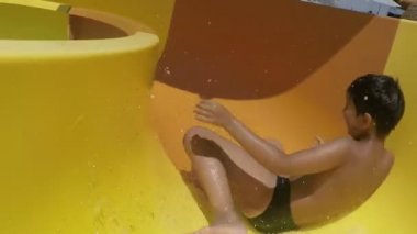 Laughing boy is rotating and riding down orange slide in a water park