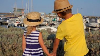 Şapkalı kız ve erkek yat limanında el ele tutuşuyorlar.