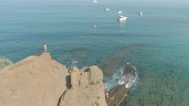 Disparos aéreos de un hombre de pie sobre un acantilado sobre un mar transparente — Vídeo de stock