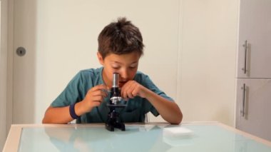 Meraklı, siyah saçlı genç bir çocuk mikroskoptan bakıyor.