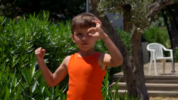 Маленький веселый мальчик в оранжевой футболке показывает свои бицепсные мышцы — стоковое видео