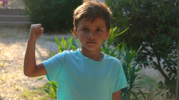 Маленький веселий танірований хлопчик у синій футболці показує його м'язи біцепсів — стокове відео