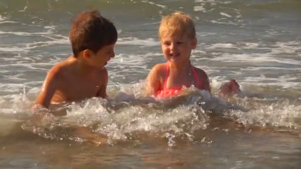 Little boy and a girl in pink swimsuits are sitting in a splashing sea waves — Stock Video