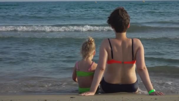 Mamá e hija están sentadas en la orilla del mar con la espalda a la cámara — Vídeos de Stock