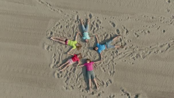 Tiro aéreo de uma equipe amigável de crianças em roupas brilhantes jazem na areia — Vídeo de Stock