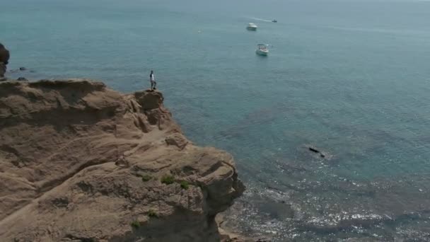 Schieten vanuit de lucht van een man die op een klif boven een heldere Middellandse Zee staat — Stockvideo