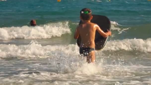 Αγόρι τρέχει μέσα στα κύματα της θάλασσας με σανίδα κολύμβησης — Αρχείο Βίντεο