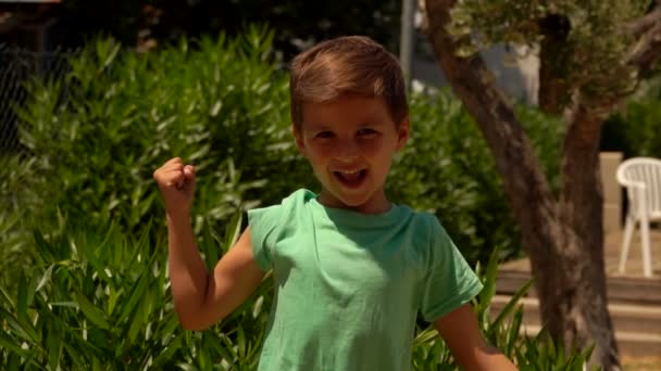 Ένα μικρό χαρούμενο αγόρι με πράσινο μπλουζάκι δείχνει τους δικέφαλους μυς του. — Αρχείο Βίντεο