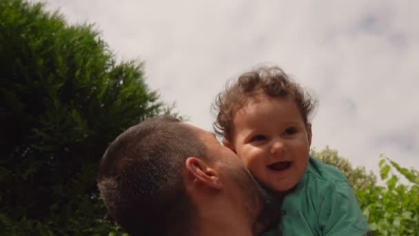 Stilig pappa leker och kysser en lockig glad pojke utomhus i trädgården — Stockvideo