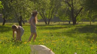 Şapkalı iki kız bahçede çiçek topluyor ve çelenk yapıyor.