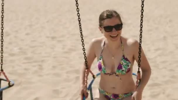 Young girl in swimsuit and sunglasses swinging on swing on the beach — Stock Video