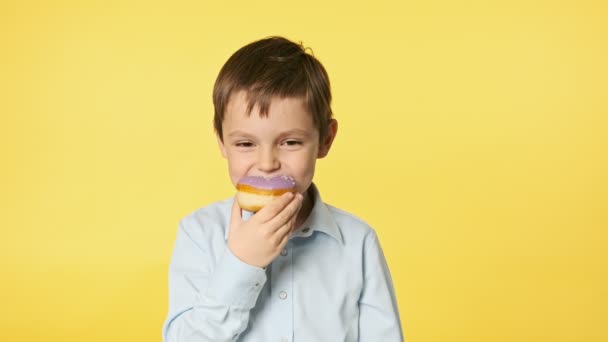 可爱的小男孩在黄色背景下吃甜甜圈 穿着蓝色衬衫的可爱的高加索男孩的特写 带着紫罗兰的光泽吃甜甜圈 味道真好 Uhd — 图库视频影像