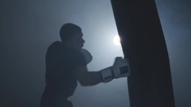 Yalnız boksör karanlık spor salonunda kum torbasına vurur. Genç adam içeride antrenman yapıyor. 4k, Uhd