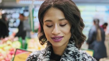 Güzel bir kadın kameraya bakıyor ve arka planda alışveriş yaparken gülümsüyor..