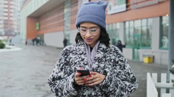 Joven mujer afroamericana en gafas usando smartphone cerca del centro comercial . — Vídeos de Stock