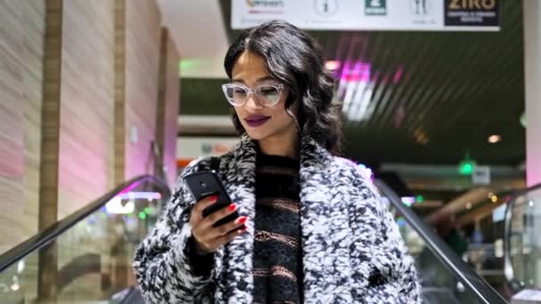 Afrikanerin mit Brille bedient Smartphone auf Rolltreppe in Einkaufszentrum — Stockvideo
