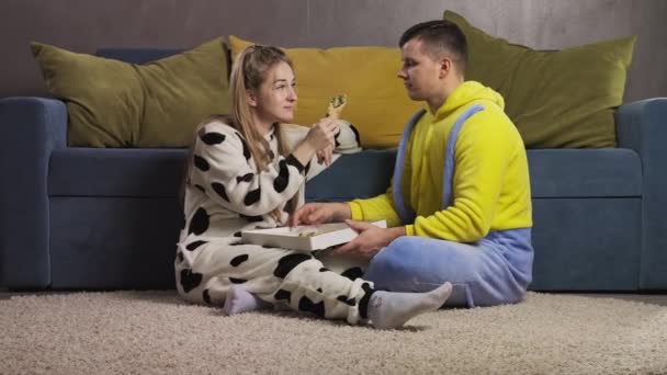 Jonge gelukkige man en vrouw zitten thuis op de vloer en eten smakelijke pizza. — Stockvideo