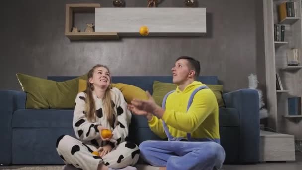 Pareja feliz en pijama divirtiéndose en la sala de estar y haciendo malabares con naranjas — Vídeo de stock